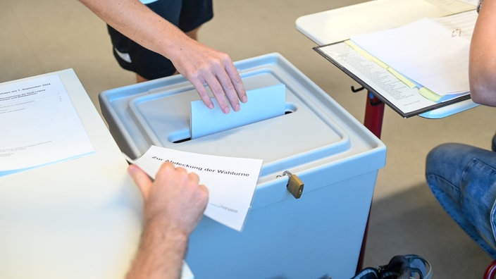 Eine Hand steckt bei einer Wahl den Stimmzettel in die Wahlurne