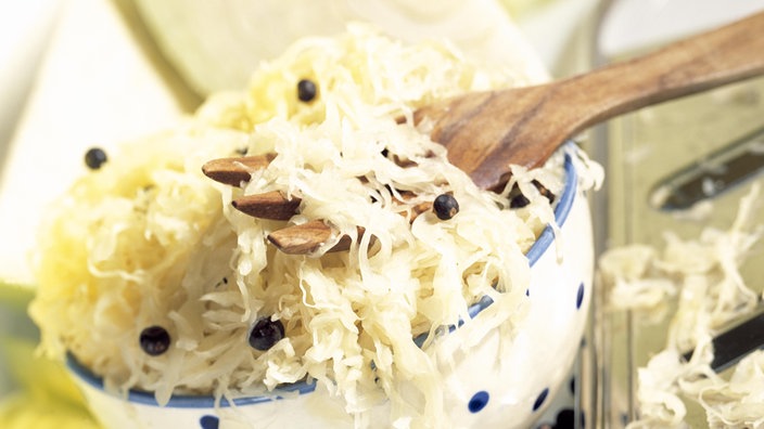 Sauerkraut mit Pfefferkörnern in einer weißen Schale mit blauen Punkten und auf einer Holzgabel.