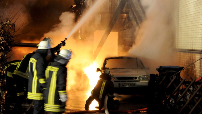 Vier Feuerwehrmänner mit weißen Helmen und Schutzkleidung versuchen mit Wasser einen Hausbrand zu löschen. Das Haus ist vor lauter Rauch und Wasserdampf kaum zu erkennen.