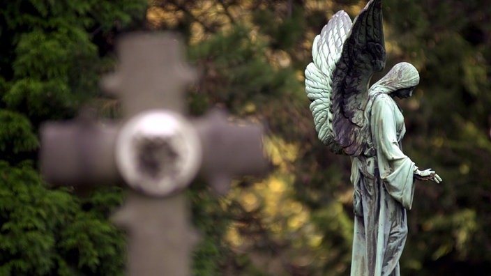 Vor grünen Bäumen und Sträuchern erkennt man schemenhaft die Umrisse eines Grabkreuzes. Rechts daneben die grün oxydierte Metallstatue eines beflügelten Engels mit gesenktem Haupt.