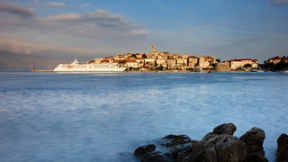 Blick übers Meer auf die historische Altstadt von Korčula, an der eine weiße Fähre vorbeifährt.