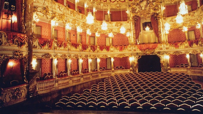 Der Rokoko-Saal des Cuvilliés-Theaters. Emporen über vier Stockwerke ermöglichen Besuchern eine gute Sicht. Sie sind mit Schnörkeln verziert und goldweiß lackiert. Eine edle Tapete ziert die Wände. Kronleuchter hängen von der hohen Decke und schaffen eine festliche Stimmung.