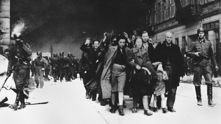 Eine Gruppe von Männern, Frauen und Kindern wird von Soldaten mit Gewehren durch eine Straße getrieben, im Hintergrund ist Rauch zu sehen.