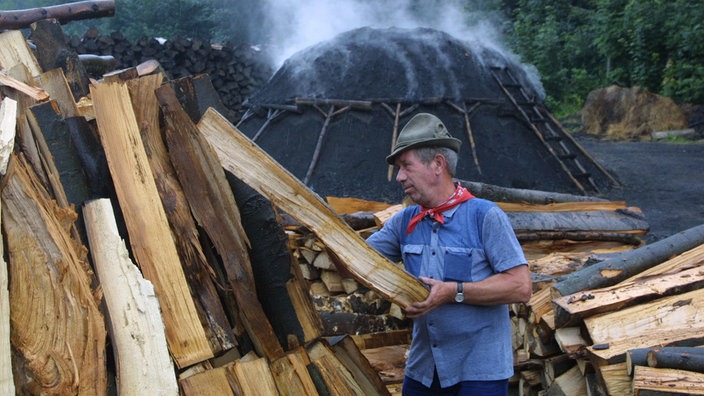 Im Vordergrund schichtet ein Köhler Holzscheite auf. Im Hintergrund ein qualmender Meiler.