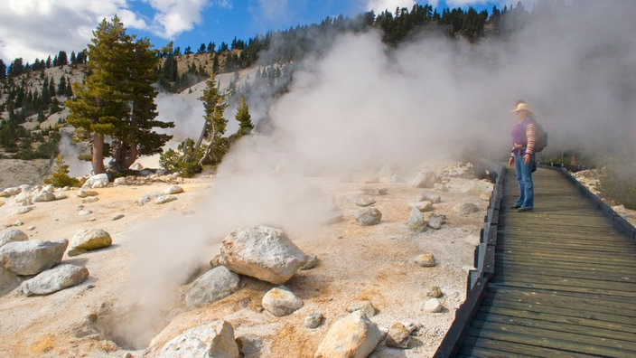 Ein Besucher des Lassen-Volcanic-Nationalparks steht auf einem hölzernen Pfad neben einem Erdloch aus dem heißer Dampf empor quillt.