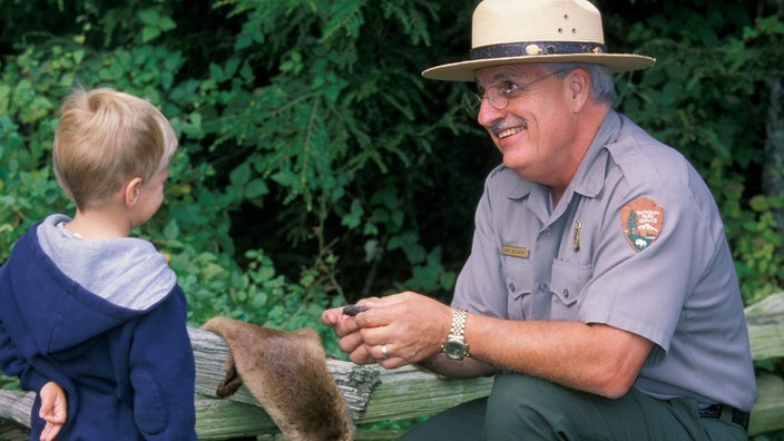 Ein älterer Mann in Ranger-Uniform hat sich neben einem kleinen Jungen in der Hocke niedergelassen und erklärt ihm etwas über ein Tierfell.