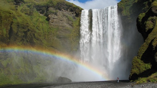 Von einem Felsen stürzt ein Wasserfall in die Tiefe. Ein Regenbogen liegt über dem fallenden Wasser.