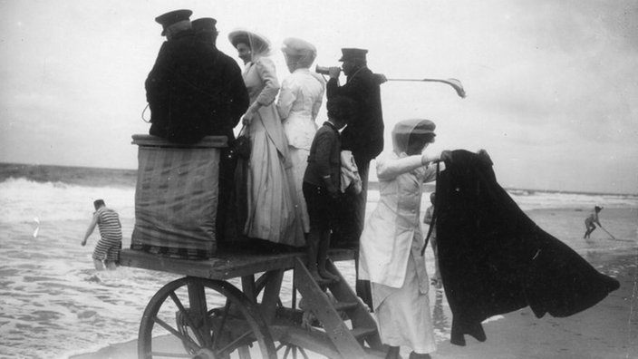 Schwarzweiß-Foto: Badewagen am Strand mit Damen in weißen Kleidern und Herren in Anzügen.