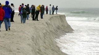 Menschen gehen im Winter am Strand spazieren und beobachten an einer Abbruchkante, wie das Meer den Sand fortspült.