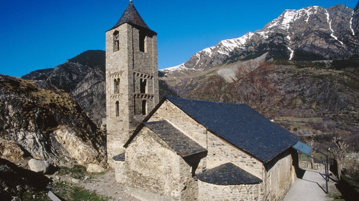 Blick auf eine alte kleine Kirche. Im Hintergrund felsige und verschneite Berge.