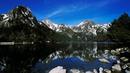 Blick auf einen See in den Pyrenäen, dahinter verschneite Berge.