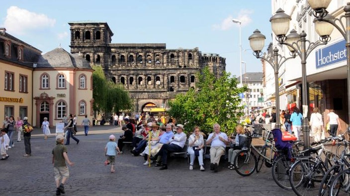 Blick von der Fußgängerzone auf die Porta Nigra. Auf der Straße spielen Kinder und bummeln Passanten. Auf einer Bank sitzen Menschen.