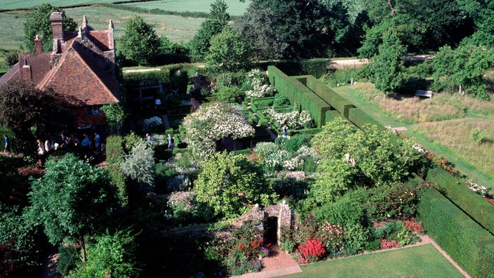 Das Bild zeigt einen Garten von oben. Es sind Bäume, Hecken und Blumenbeete zu sehen. Auf der linken Seite steht ein kleines Haus.