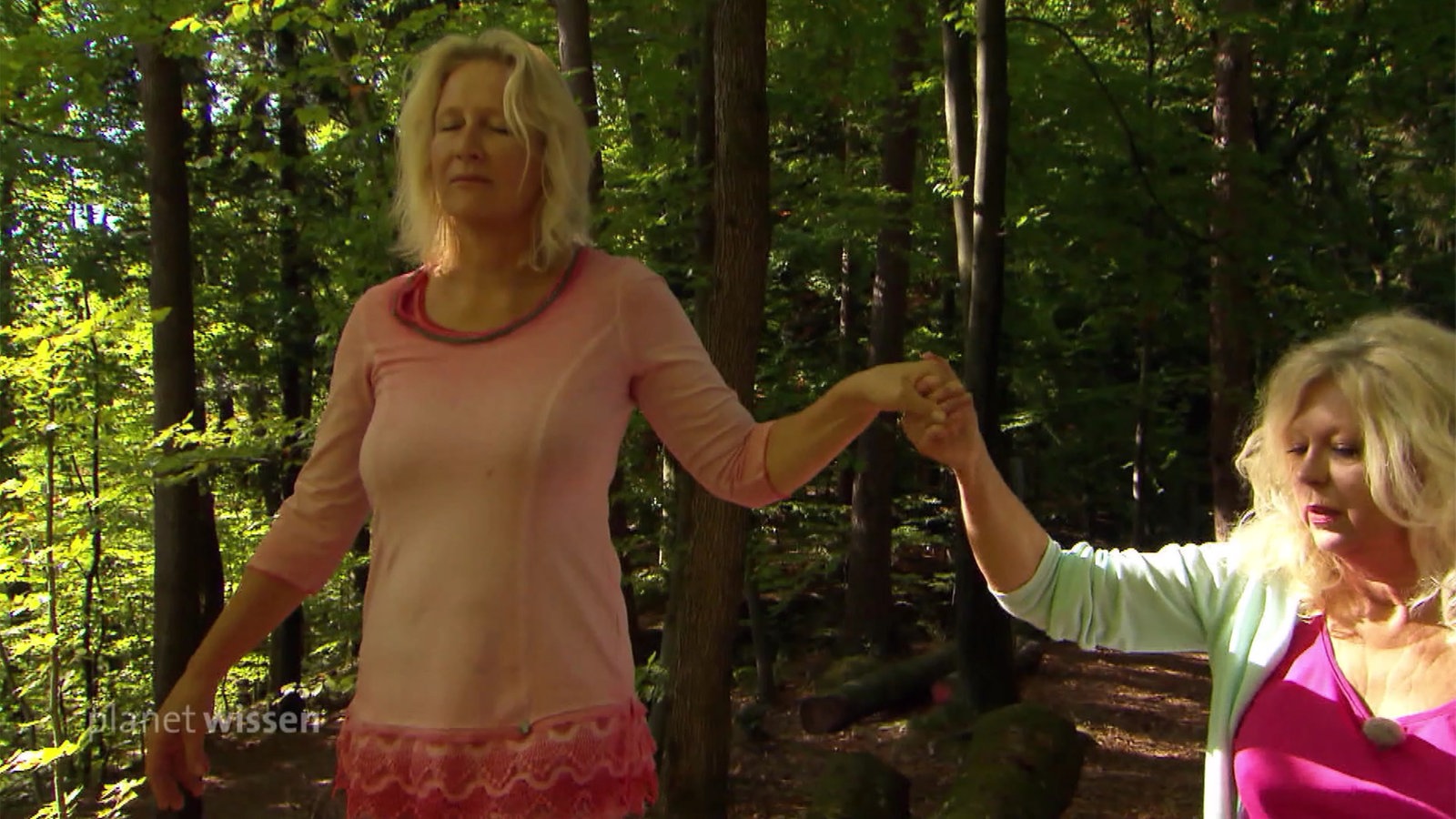 Eine Frau lässt sich mit geschlossenen Augen von einer anderen Frau an der Hand durch den Wald führen.